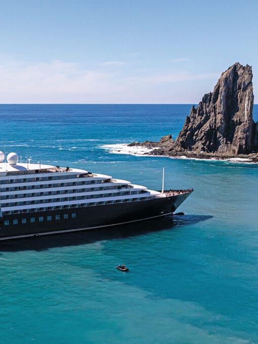 Large black and white cruise ship in a light blue sea with a rock sculpture behind it.