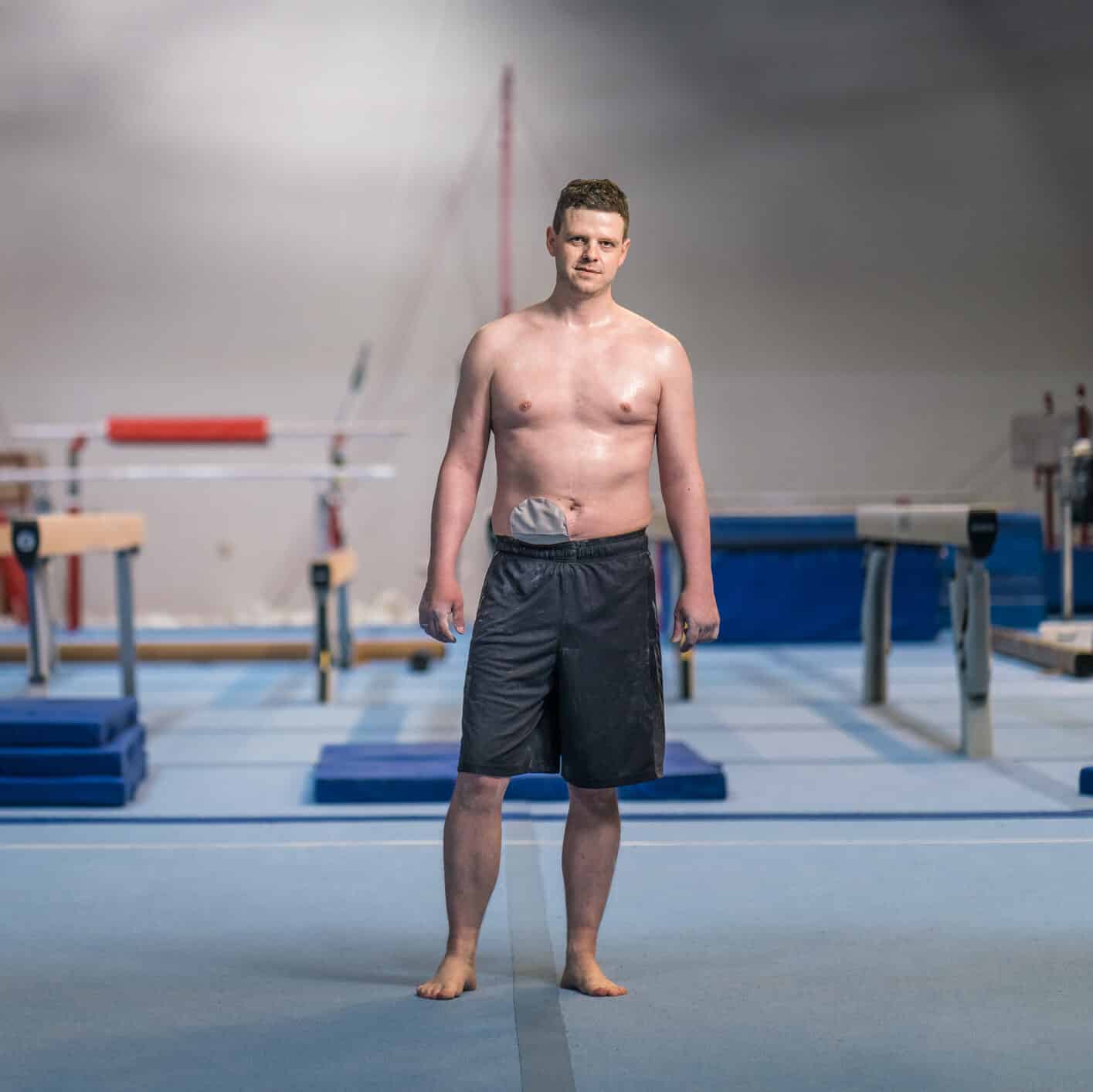 Full length of shirtless male athlete with stoma bag standing at gym. Portrait of confident sportsman is against gymnastics equipment. He is at health club.
