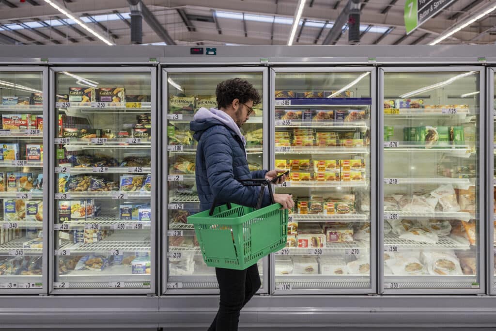 Man shopping in a supermarket while on a budget. He is looking for low prices due to inflation, standing looking at his phone in front of a row of freezers. 