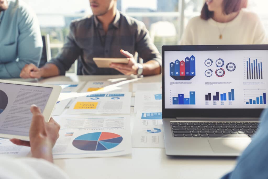 Group of people meeting with technology and paperwork. Close up of digital tablet and laptop computer with market analysis data on the screen. The documents have financial or marketing figures, graphs and charts on them. Multi ethnic group including Caucasian, latin and African American