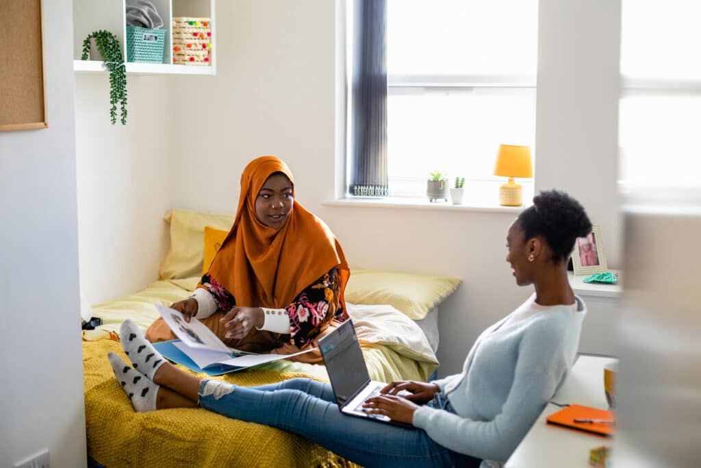 Two female uni student friends sitting in their dorm bedroom are studying together while sitting on the bed in the North East of England. They are using laptops/have their work files in front of them.